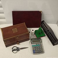 Photo showing checkbook binder, wooden desk box with brass accents, calculator, black hole puncher, scissors, and small desk accessories arranged on a white background.