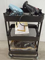 Photo showing 2 hairdryers on top shelf of black metal utility cart with wheels; also includes boxed new mirror on middle shelf and packaged wig on bottom shelf.