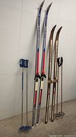 Full view of three skis and two pairs of ski poles standing upright against wall