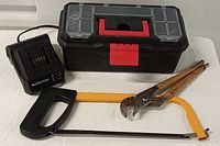 Photo showing the black toolbox with red latch, transparent compartments, yellow and black hacksaw, rusted locking pliers, and Black & Decker battery charger.