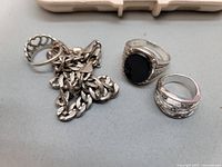 Silver chain bracelet with winged figure pendant, silver ring with black oval stone, wide silver ring on grey background