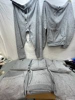 Front view of one grey Champion kids medium track pants and zip hoodie set hung on wall with four other folded sets below on table.