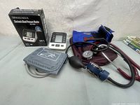 Photo showing two blood pressure testers: a DINGGUAGUA electronic arm blood pressure monitor with its box and cuff, and a manual sphygmomanometer with blue cuff and stethoscope.