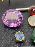 Close-up photo showing large faceted purple gemstone and small blue gemstone set in gold-toned metal.