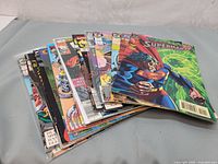 Photo showing a stack of approximately 15 various comic books fanned out, featuring colorful covers with superheroes and other characters.