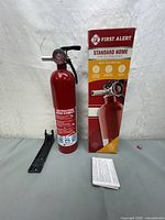Photo showing the fire extinguisher standing beside its original red and yellow box with instructions and mounting bracket laid out.