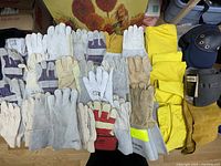 Photo showing multiple pairs of work gloves in white, beige, tan, and leather, yellow rain suits, and black knee pads