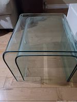 Pair of two transparent curved tempered glass end tables placed on a wooden floor, showing their size and shape.
