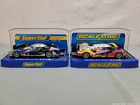 Two 1:32 scale slot car models in display cases side by side, one from SuperSlot and the other Scalextric.