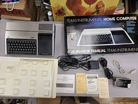 View of the TI-99/4A computer next to original box, with power supply, RF modulator, cables, and manuals arranged around the device
