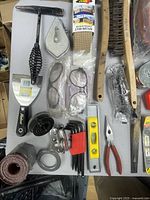 Photo showing assorted hand tools and accessories laid out on a table surface including pliers, Allen wrenches, level, brushes, safety glasses, and a putty knife.
