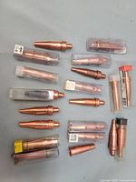 Top-down view of multiple copper torch welding tips, various shapes and sizes, several in plastic protective tubes, some loose on a grey surface.
