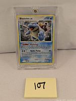 Front angled view of Blastoise LV.52 Pokemon card in protective case with yellow sticky note labeled '107'