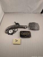 Photo showing the three items: a pistol-shaped thermometer, Gannon DynaBuckle belt buckle, and National Final Rodeo Hesston belt buckle.