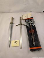 Mini decorative swords and double headed axe displayed on white fabric background showing details of blades and handles.