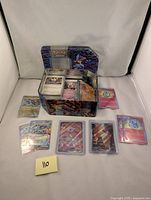 Wide shot of multiple Japanese Pokemon cards including holo and reverse holo cards placed around a metal Pokemon tin, showing condition and storage.