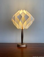 Front view of the lamp turned on, showing the glow of the sculptural white plastic shade and the wooden and metallic base.