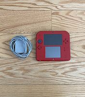 Red Nintendo 2DS handheld console with charger cable and plug laid on wooden floor.