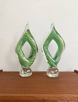 Front view of two matching green swirled art glass sculptures resting on a wooden surface against a plain white wall.