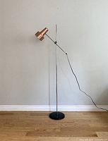 Full view of the floor lamp against a plain wall, showing the copper shade, chrome pole, black base, and power cord.