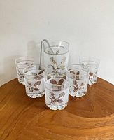 Front view of vintage Italian frosted glass ice bucket with gold leaf pattern and six matching drinking glasses arranged around it on wooden surface.