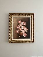 Framed oil painting showing pink roses in brown vase against dark background, hanging on a wall.