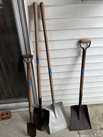 Photo shows four assorted shovels arranged vertically against a white siding wall. Two have round metal blades, one is a flat silver blade, and one is a square dark blade with D-handle.