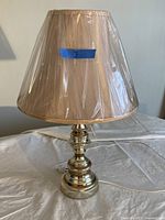 Brass table lamp with beige fabric shade covered in protective plastic, placed on a white tablecloth.