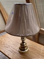 Full view of brass table lamp with factory-covered lampshade on wooden surface near windows with blinds.