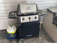 Broil King gas BBQ grill with three control knobs on the front, closed lid, and one propane tank in a blue crate beside it.
