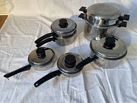 Top-down and angled view of the six-piece stainless steel cookware set, showing lids and black handles on varied sized pots and pans, including a pressure cooker.