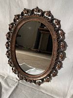 Oval decorative mirror with bronze ornate floral frame shown hanging against a light colored backdrop.