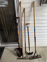 Photo showing five garden tools standing upright against white siding: shovel with D-grip handle, long-handled rake, spade, hoe, and dethatcher, all with wooden handles and metal heads.