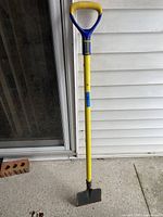 Photo of an ice chopper tool standing upright against a white siding wall and a sliding glass door. The tool features a yellow metal shaft, a blue metal structural support near the handle, a wooden hand grip, and a flat metal blade at the bottom.