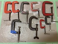 Photo showing 11 various sized and colored metal C clamps arranged on floor.