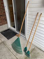 Photo showing three lawn rakes leaning against a porch wall and door, including one large green plastic fan rake and two smaller fan rakes with wooden handles in good condition.