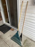 Two lawn rakes leaning against the exterior siding of a house, showing their full length and details of the rake heads and wooden handles.