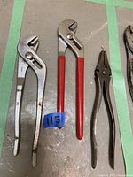 Three adjustable pliers laid out on a grey surface. Two are silver with one having red handles, the other plain metal. The third is smaller and brown with serrated jaws.