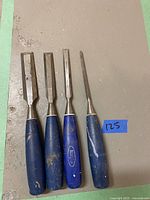 Four wood chisels with blue handles laid out on a beige surface. Each chisel has a metal blade and a blue handle showing some wear and dirt.