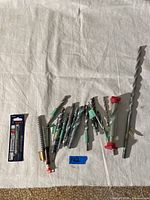 Photo shows a collection of assorted concrete drill bits and accessories arranged on light fabric background. Includes a few wrapped in tape or in plastic tubes, and one sealed in original packaging.