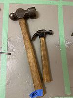 Photo showing two hammers placed side by side on a surface. The larger hammer is a ball pein hammer with a worn wooden handle and a rounded metal head. The smaller hammer is a claw hammer also with a wooden handle and metal head.