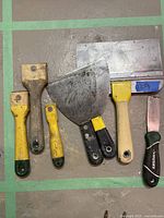 Seven paint scrapers and drywall knives arranged side by side on a floor with a green tape grid.