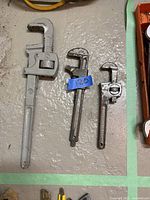 Photo showing three plumbers wrenches arranged on floor, one large silver wrench and two smaller darker wrenches.