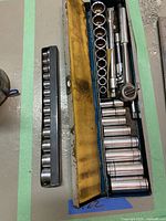 Photo of an open blue metal tool case containing various sockets, two socket extensions, and a ratchet wrench, alongside a separate socket holder strip with multiple sockets neatly arranged.