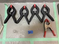 Five spring clamps arranged on a floor including four large black clamps with red tips and one smaller silver clamp with red grips.