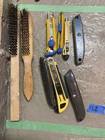 Photo showing two wooden-handled wire brushes and five utility cutting knives in various sizes and colors arranged on a concrete surface.