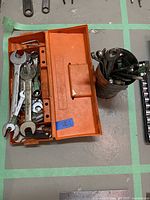 Orange metal toolbox opened showing various wrenches and tools inside. Metal container filled with additional tools next to it.