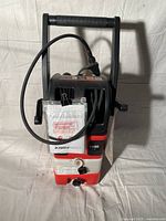Front view of the red and black electric pressure washer showing hose, water outlet, control knobs, and instruction labels.
