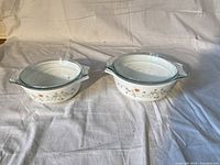 Two white glass Pyrex casserole dishes with floral decoration, each with a clear glass lid, displayed on a white fabric background.