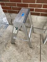 Small aluminum step ladder shown outdoors on concrete ground beside a brick wall, displaying its side profile with two steps and rubber grip top.